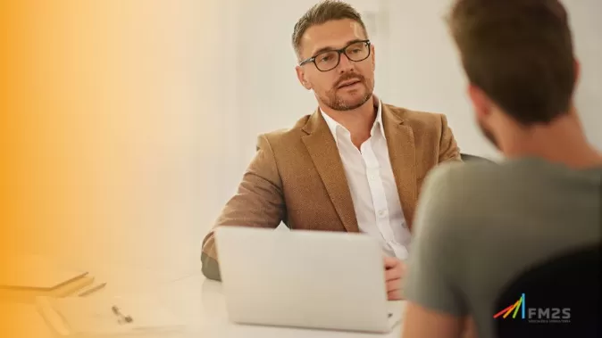 Fundo branco com degradê laranja à esquerda. À direita, um homem de terno marrom e óculos de grau sentado com um notebook em cima da mesa. No canto inferior direito, o logo da FM2S.